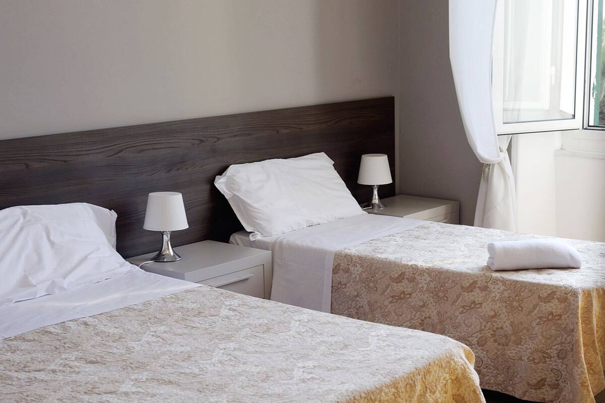 A hotel room with two single beds, each with a white pillow and a lamp.