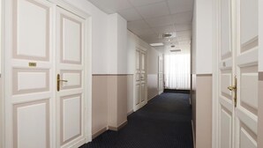 Un pasillo con puertas blancas, cada una con un número. El pasillo tiene un suelo alfombrado oscuro y paredes blancas con detalles color beige.