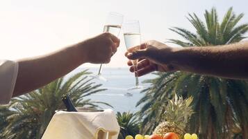 Two people toasting with champagne glasses, a fruit basket, and a bottle of wine on a table with a view of palm trees and the sea.