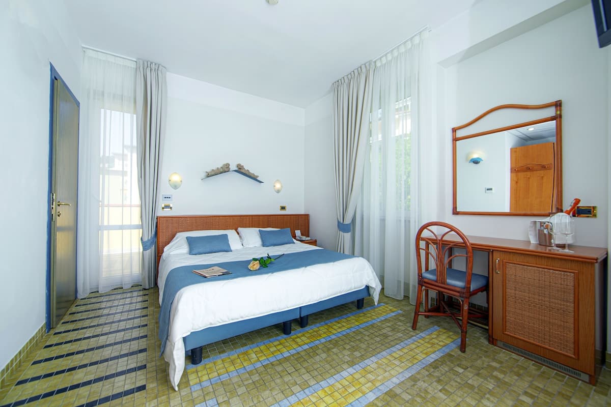A hotel room with a bed, a desk, a chair, a mirror, and a window with curtains.