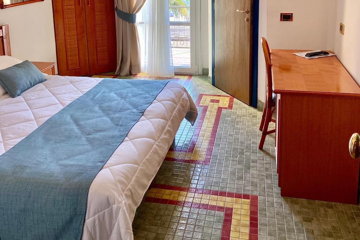 A hotel room with a bed, a desk, a chair, a wardrobe, and a view of the outside.