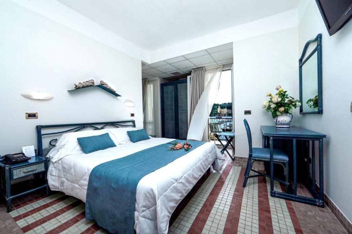 A hotel room with a bed, a desk, a chair, a mirror, and a view of the outside.