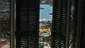 Un balcón con contraventanas abiertas, una mesa con frutero y bebidas, y vista a un puerto con barcos.