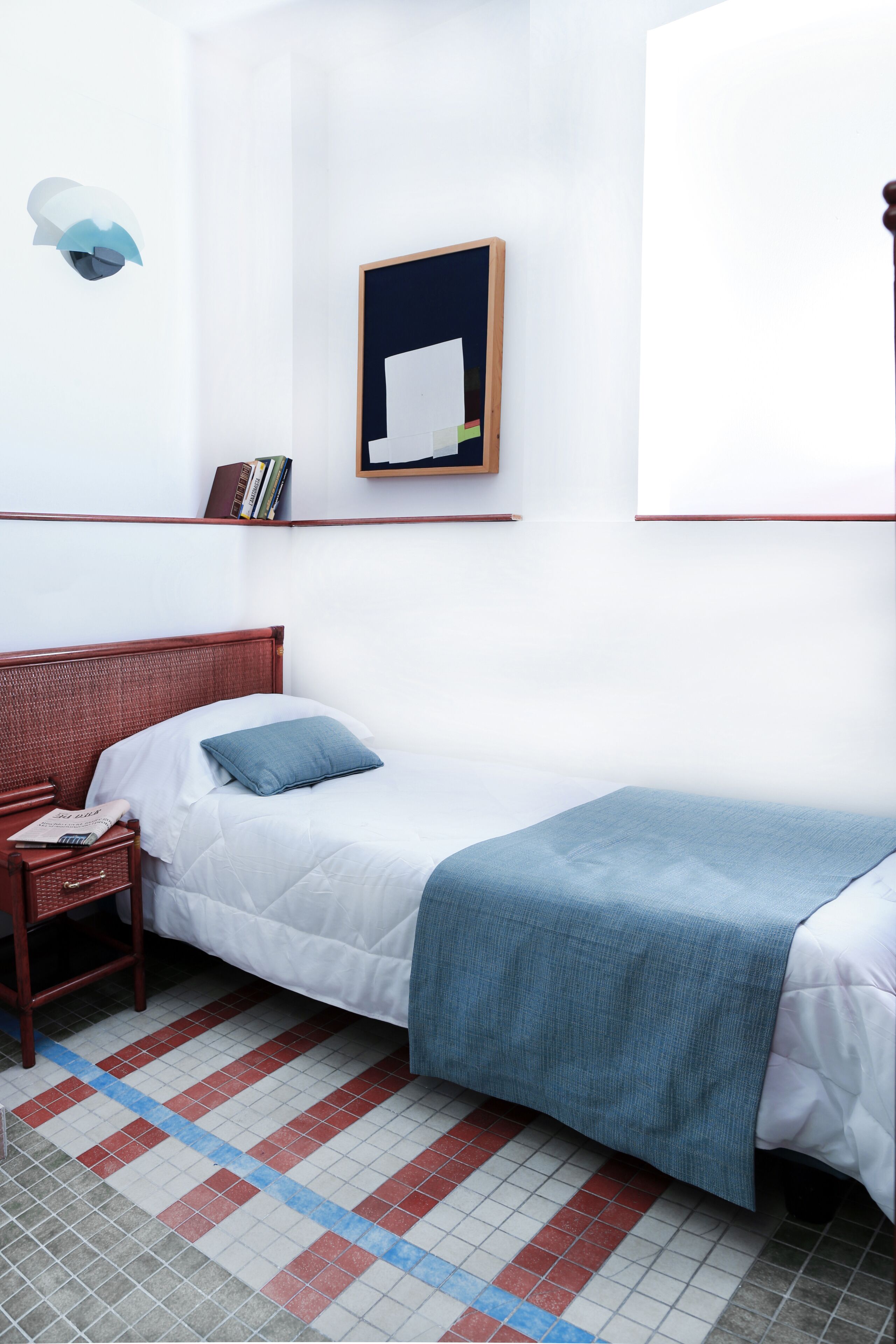 Une chambre à coucher bien rangée, avec un lit simple, une table de chevet, une commode, une étagère murale avec des livres et un tableau encadré.