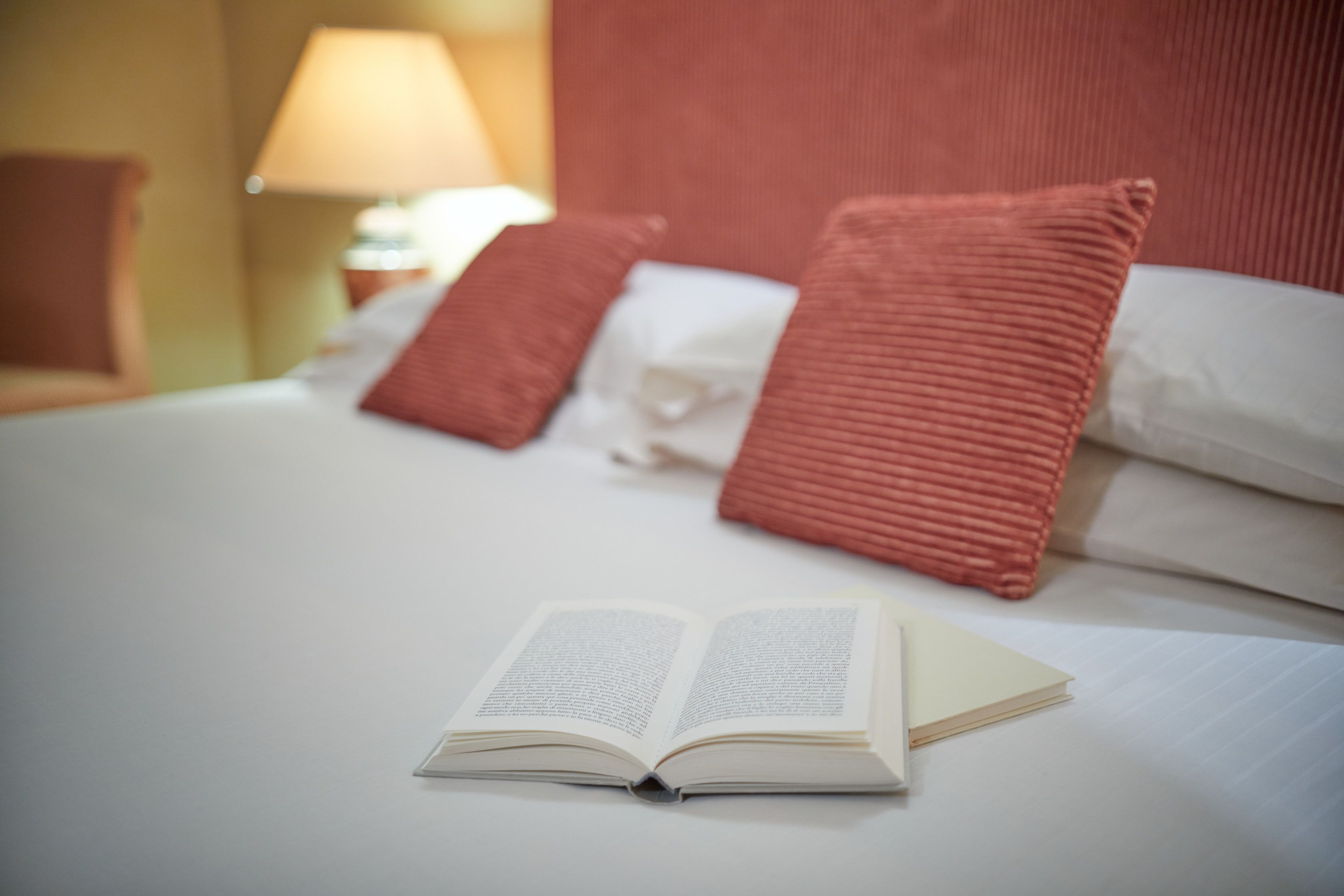 A neatly made bed with a book open on it, a lamp, and a red pillow.