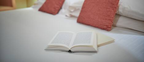 A neatly made bed with a book open on it, a lamp, and a red pillow.