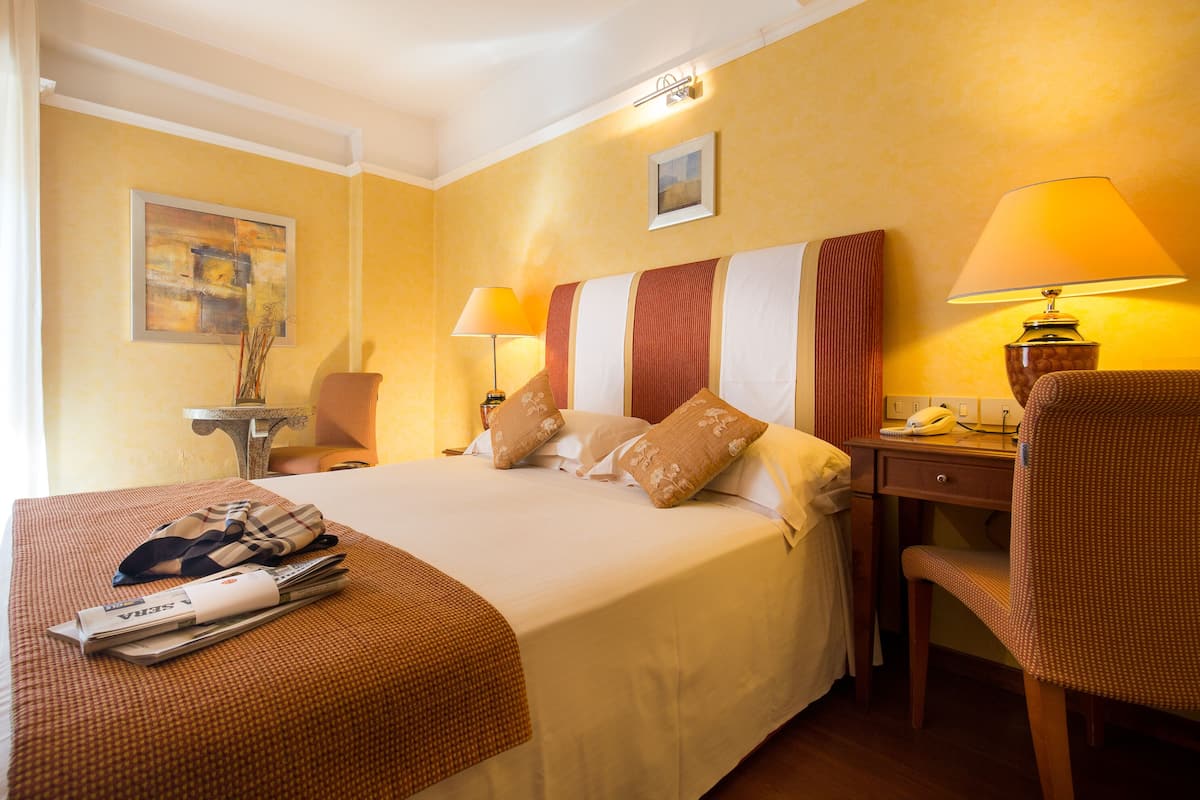 Una habitación de hotel con una cama grande, un escritorio con lámpara, una silla y una mesita con lámpara.