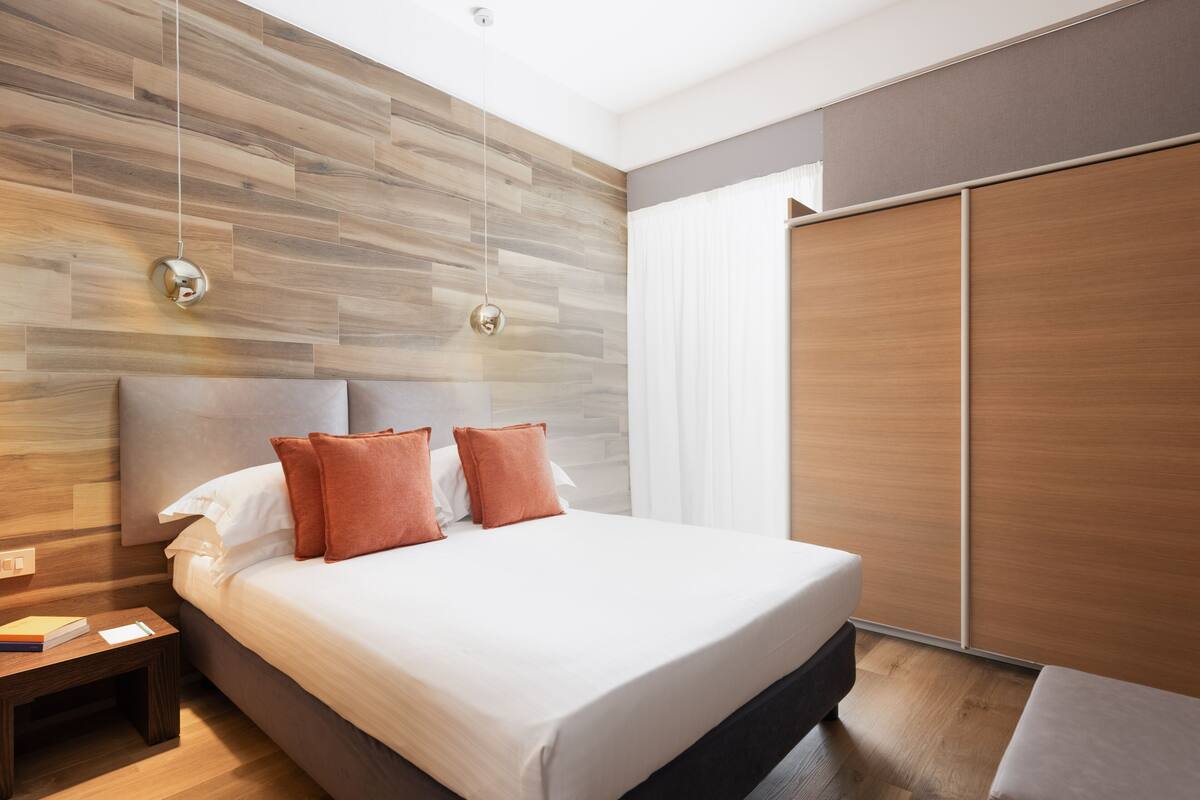 Un dormitorio moderno con cabecera de madera, una cama grande con sábanas blancas y dos almohadas naranjas, una mesita de noche con un libro y un bloc, y una puerta corredera de madera.