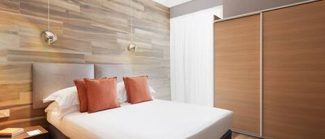 A modern bedroom with a wooden headboard, a large bed with white linens and two orange pillows, a bedside table with a book and a notepad, and a sliding wooden door.