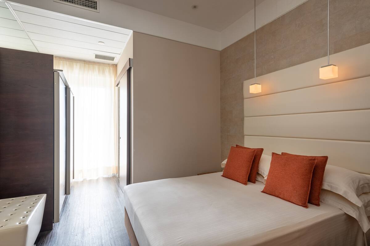 Habitación de hotel con una cama, dos almohadas, una mesita de noche y un ventanal con cortinas.
