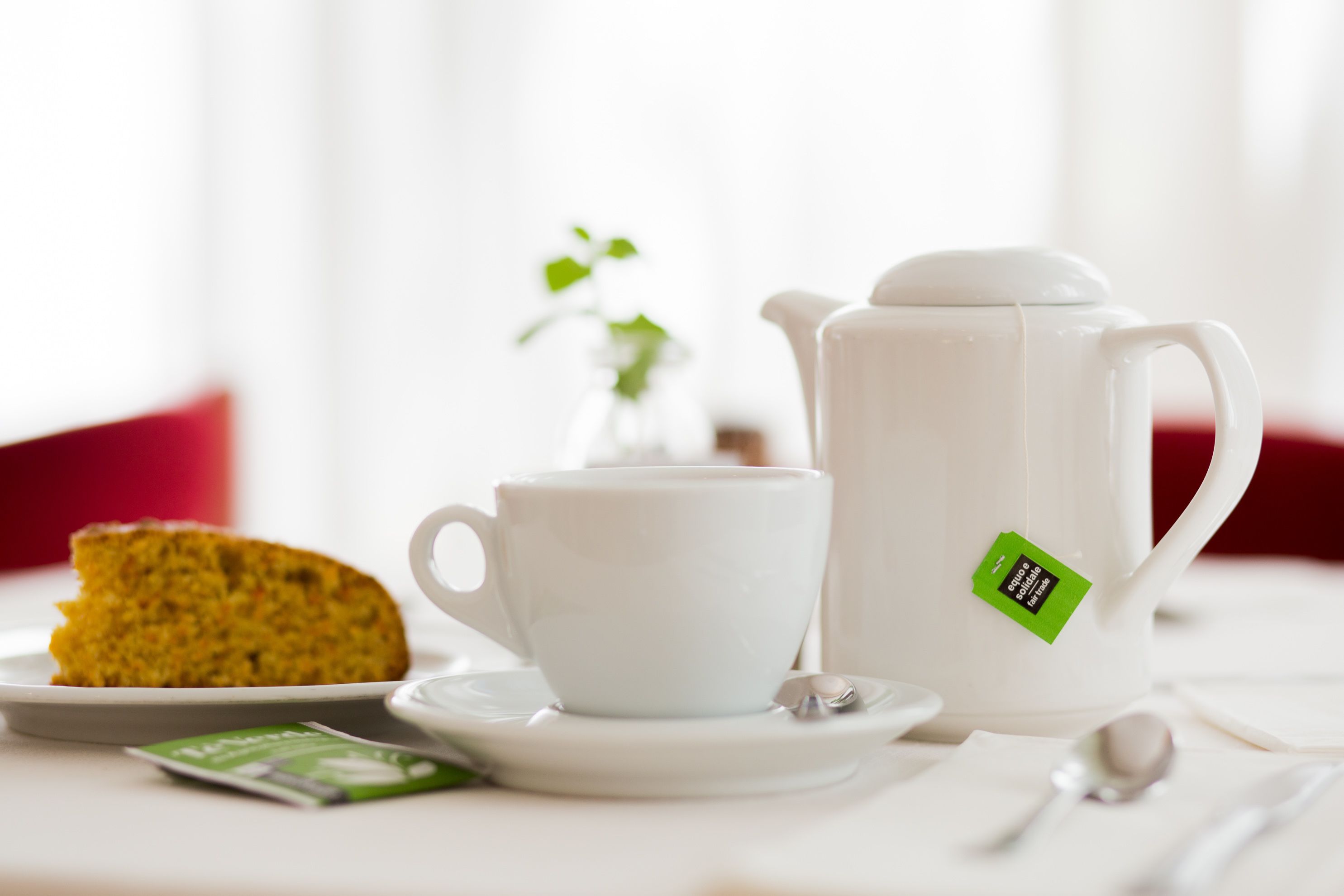 A white teapot with a green tea label, a white cup, a slice of cake, and a spoon on a white table.