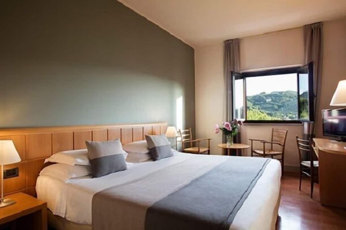 A hotel room with a large bed, bedside tables, a small table with chairs, and a window with a view of hills.