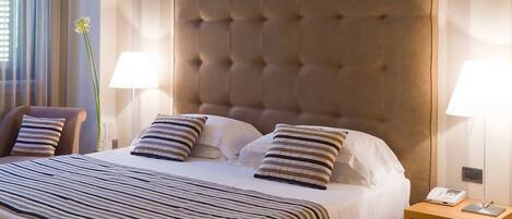 Quarto de hotel com uma cama grande, roupa de cama listrada, cabeceira estofada, duas luminárias de cabeceira e telefone ao lado da cama.