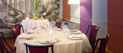Un comedor con una mesa redonda dispuesta para cuatro personas, sillas tapizadas en rojo, un jarrón con flores y un mural en la pared.