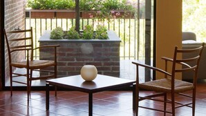 A balcony with two wooden chairs, a small table, and a vase. There is a brick wall and a window with a view of greenery and flowers. - Hotel Dei Duchi (Spoleto)
