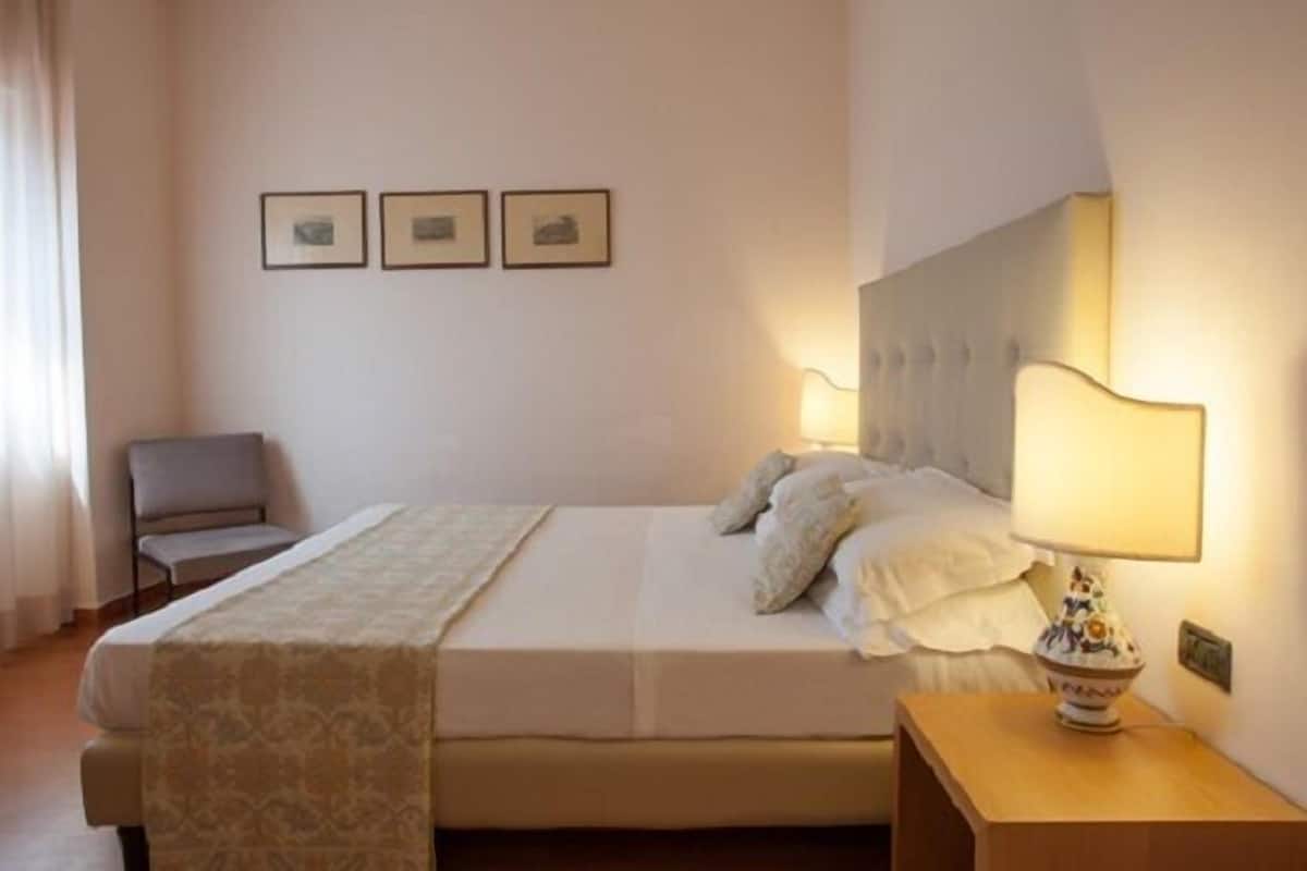 A hotel room with a bed, a bedside table with a lamp, a chair, and three framed pictures on the wall.