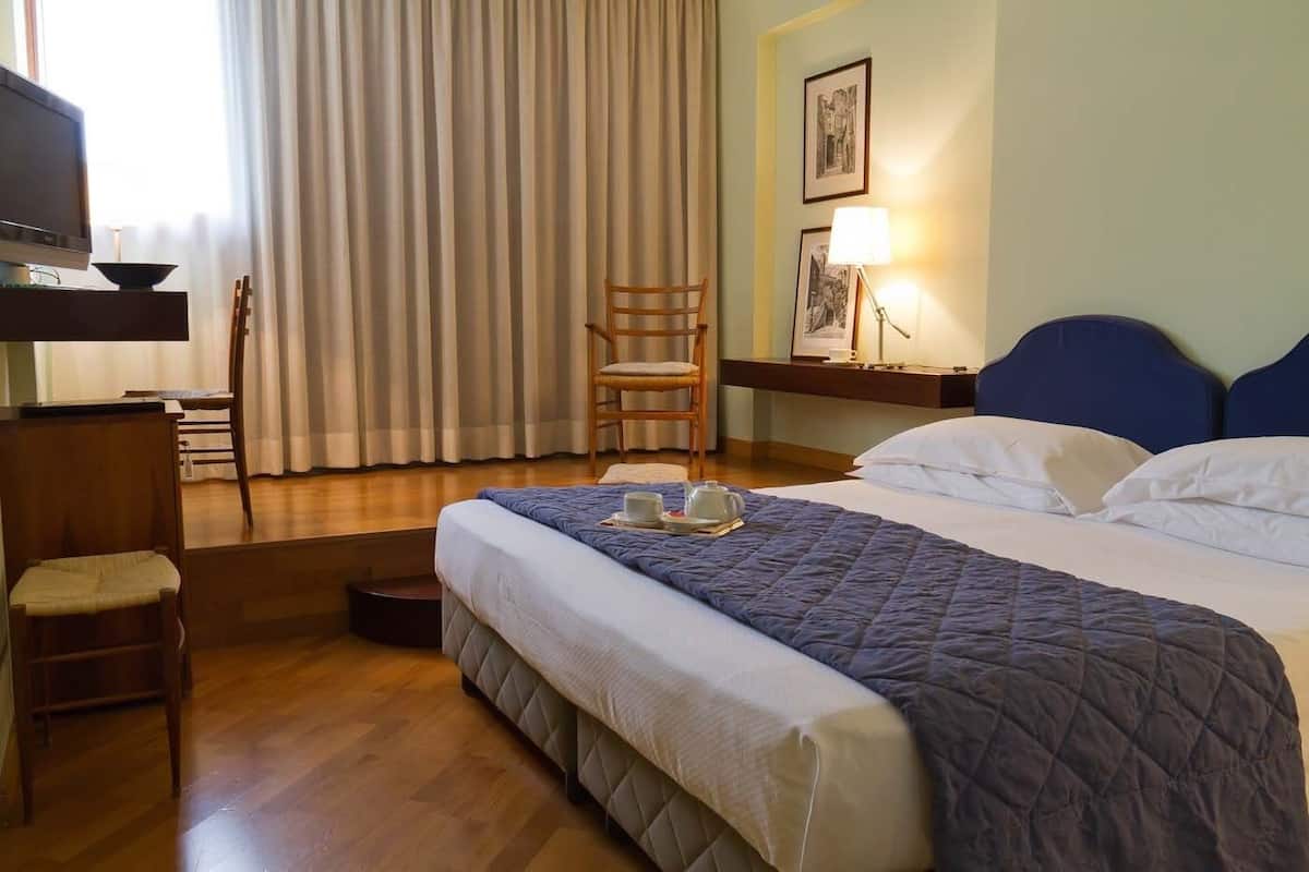 A hotel room with a bed, a desk, a chair, a lamp, and a window with curtains.