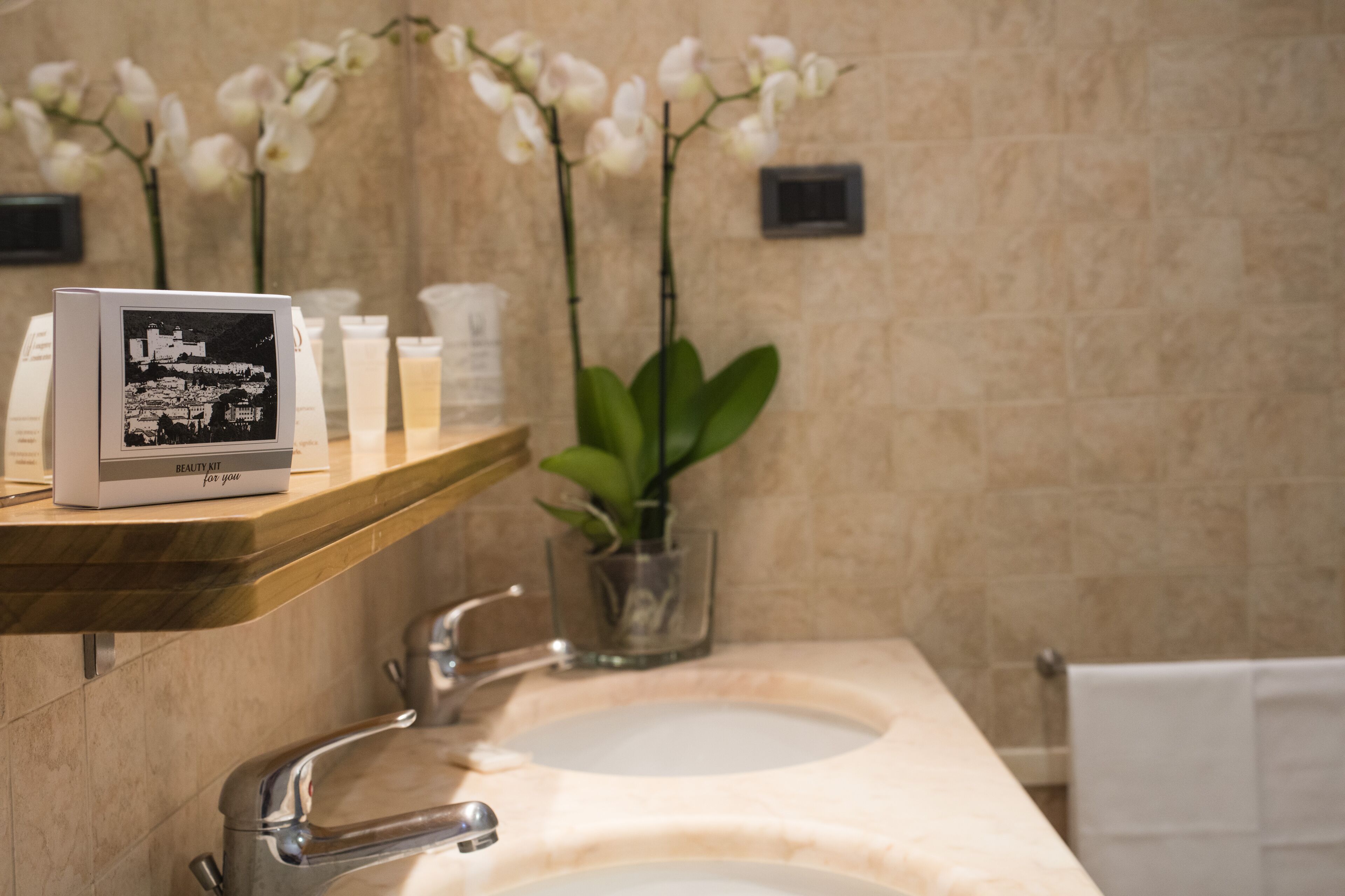 A bathroom with a marble sink, a mirror, a shelf with a beauty kit, and white orchids.