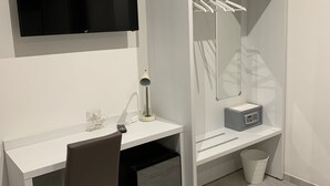 Una habitación moderna con un escritorio blanco, una silla, un televisor montado en la pared, un armario con espejo y un pequeño cubo de basura.