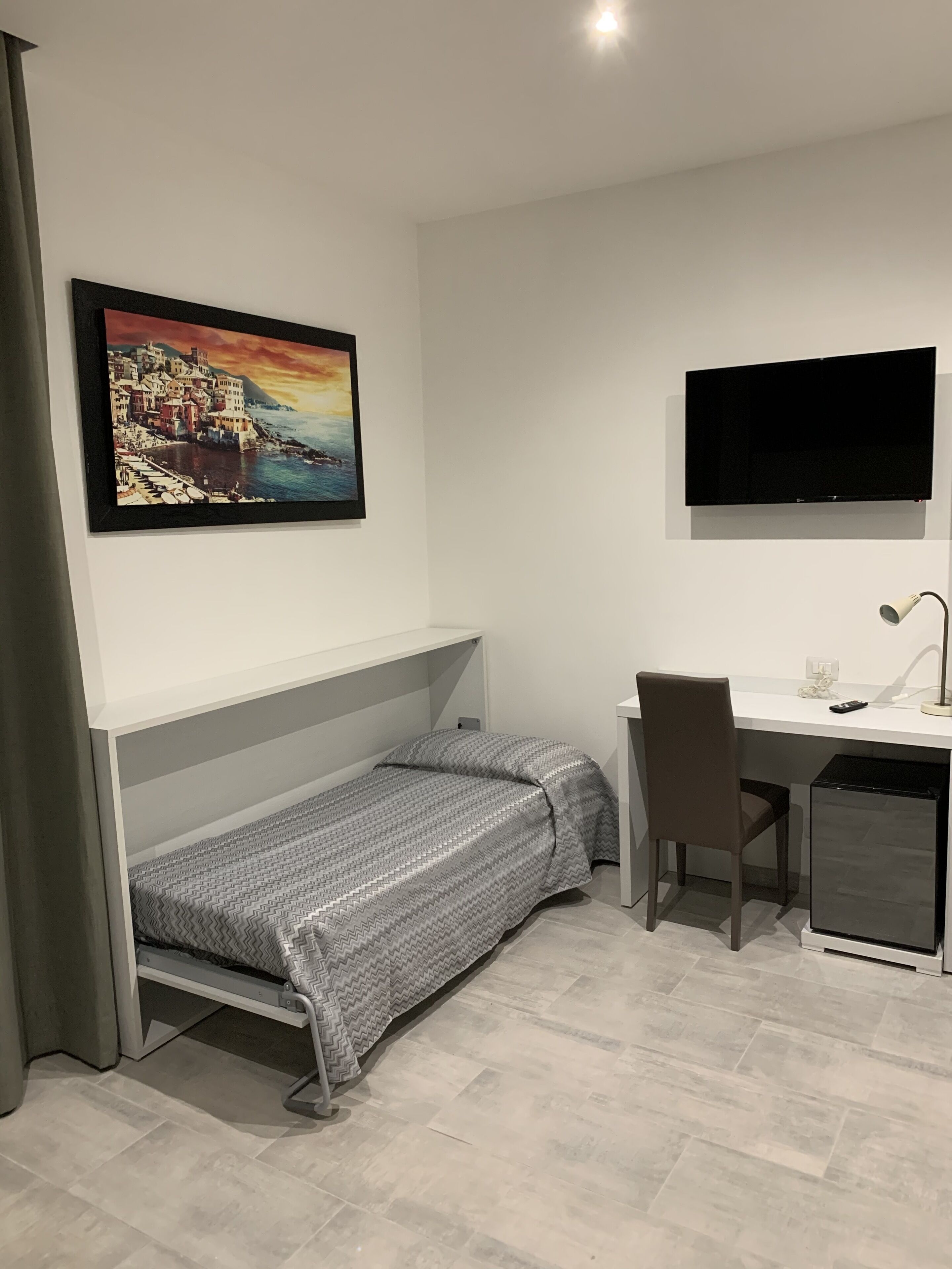 Una camera compatta con un letto, una scrivania, una sedia, una TV e un piccolo armadietto.