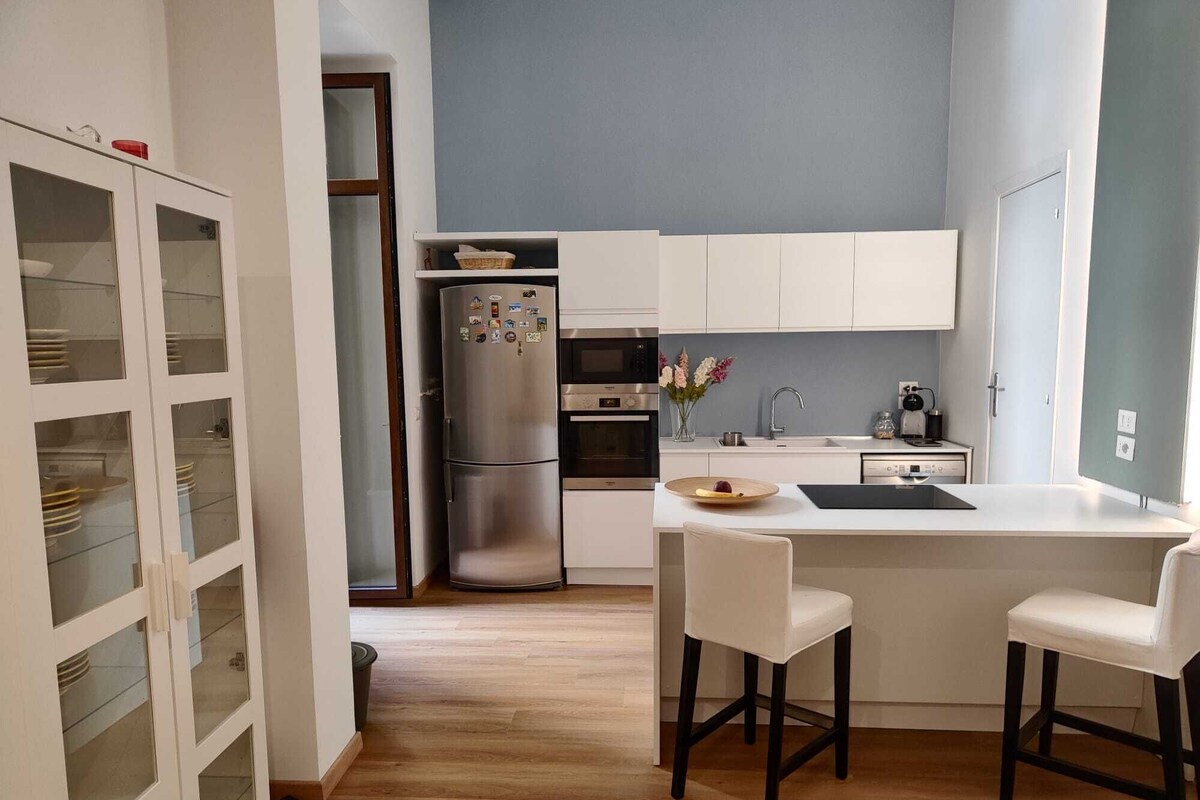 A modern kitchen with white cabinets, a stainless steel refrigerator, a built-in oven, and a small island with bar stools.