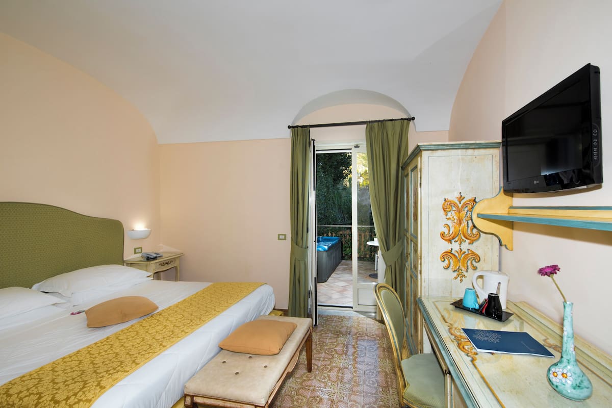 A hotel room with a bed, a desk with a vase, a television, and a view of the outside.