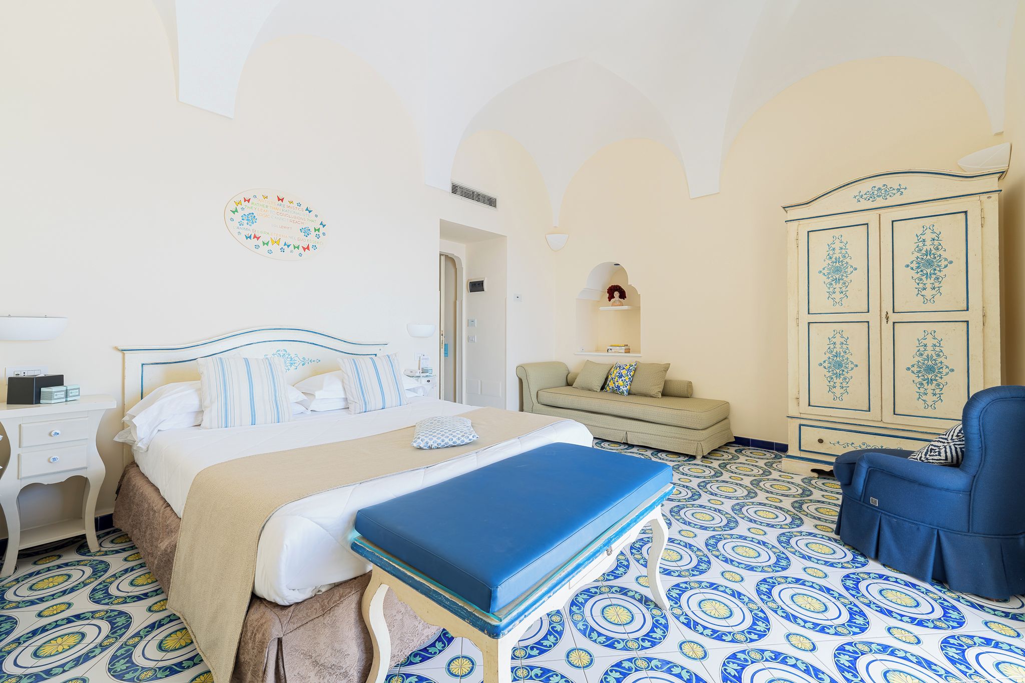 A hotel room with a bed, a sofa, a chair, a wardrobe, and a decorative wall piece.