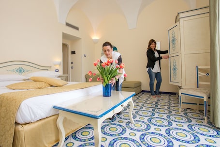 A hotel room with a bed, a table with a vase of flowers, a wardrobe, and patterned floor tiles.