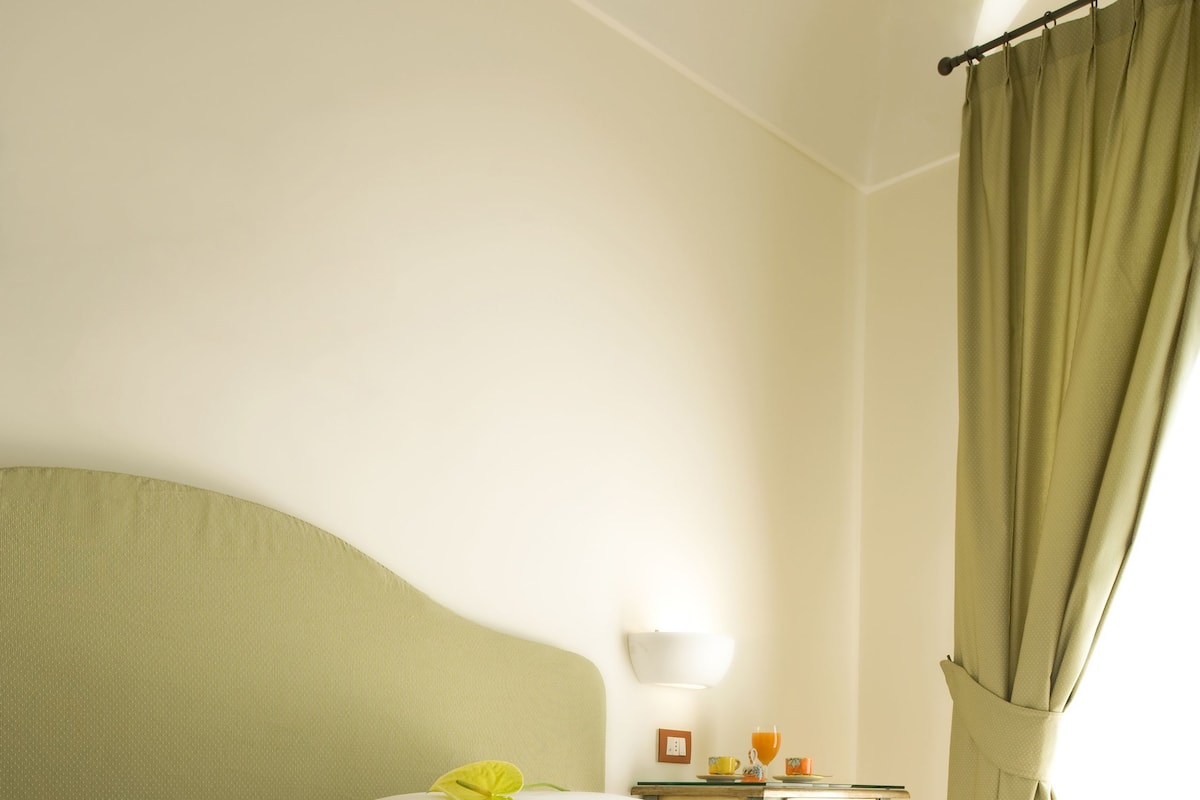 A hotel room with a neatly made bed, a small bedside table with a lamp, and a view of the outside through a window with curtains.