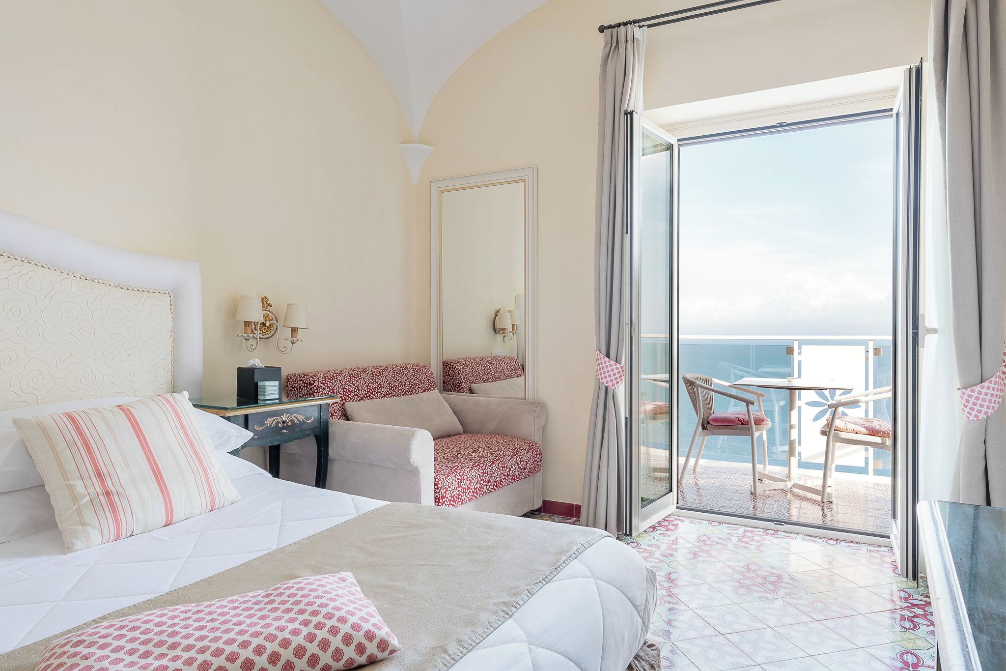 A hotel room with a bed, a desk, a chair, a sofa, and a balcony with a view of the sea.