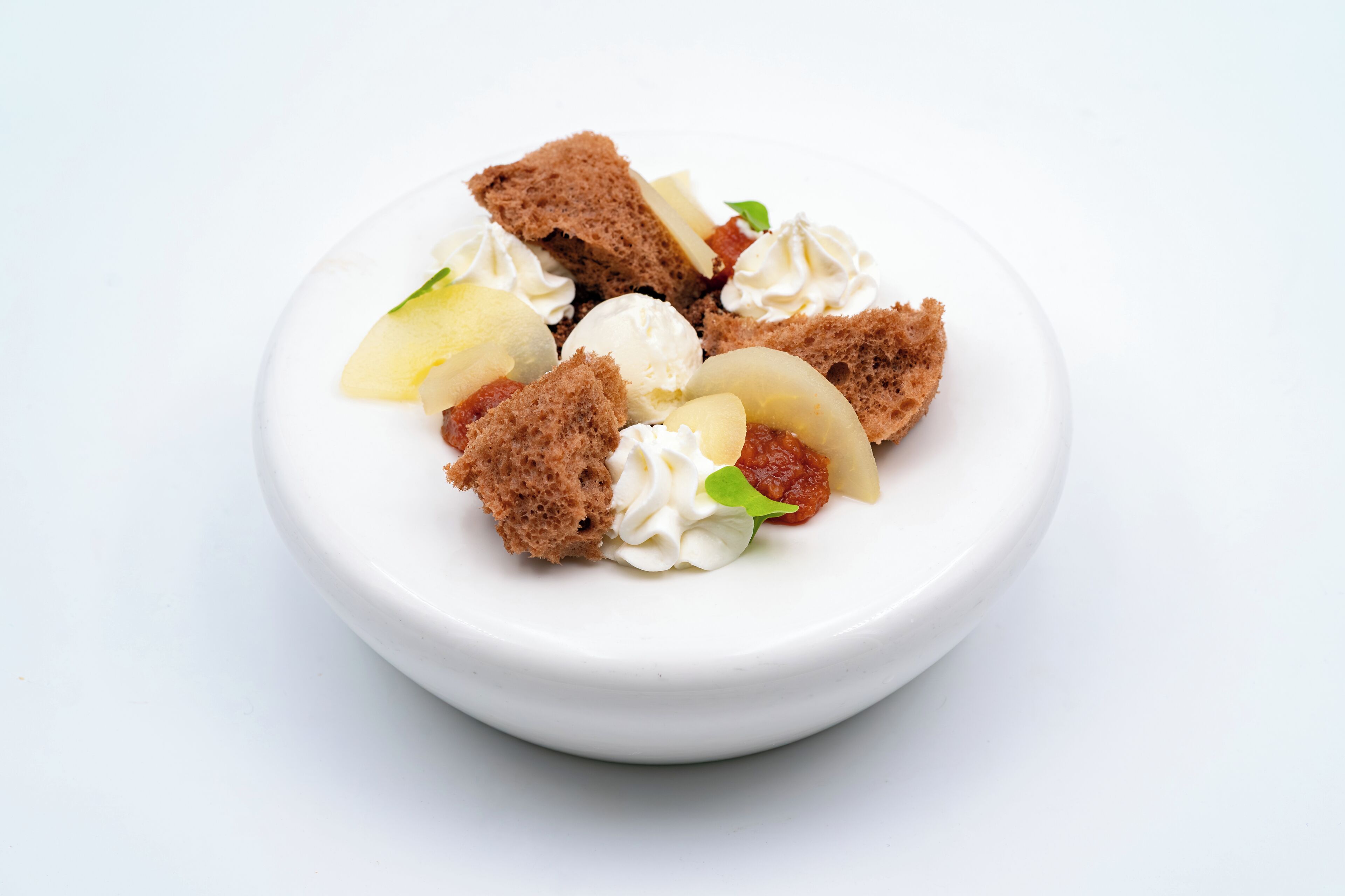 A white bowl containing a dessert with pieces of cake, cream, and fruit.