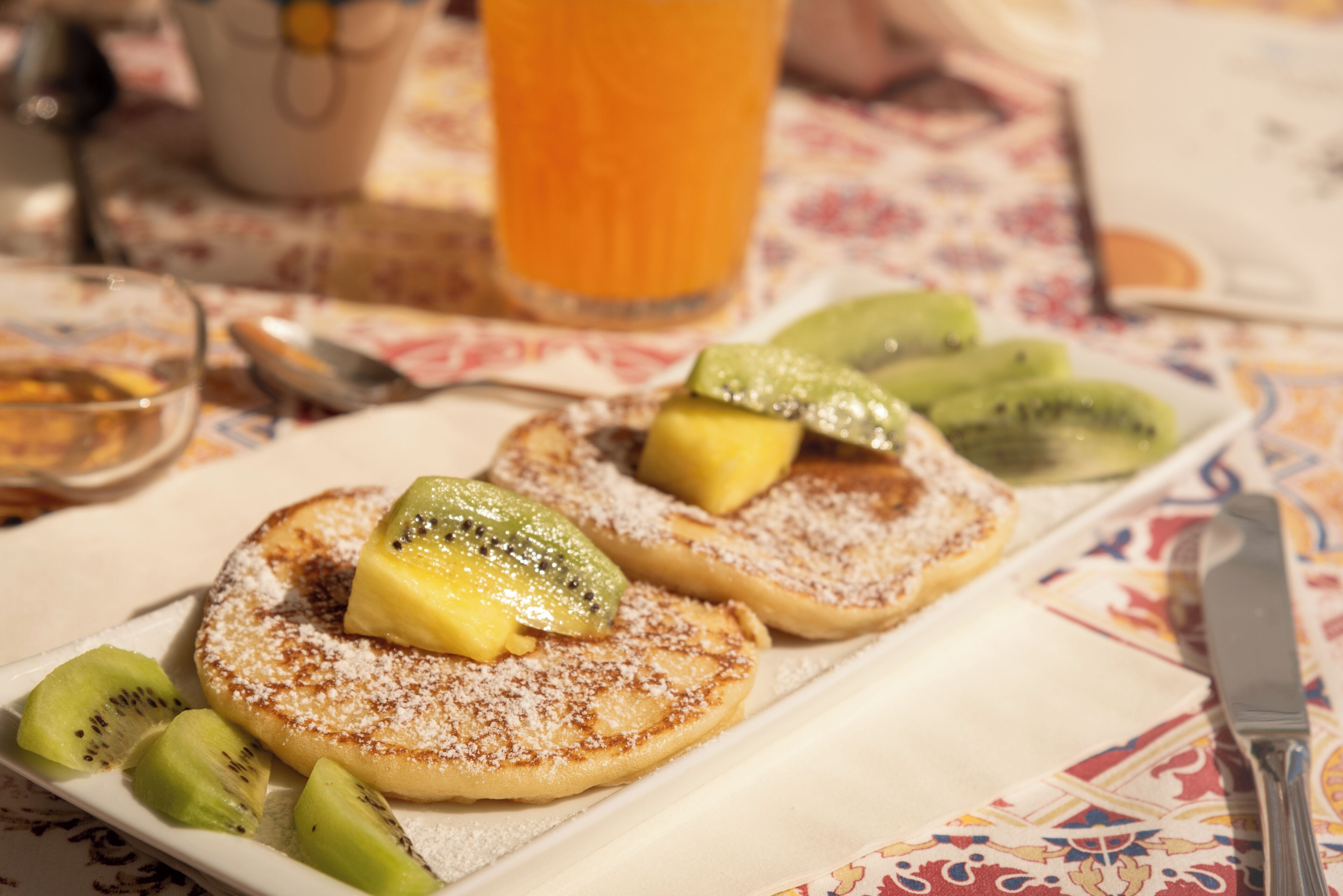 A breakfast plate with pancakes topped with kiwi slices and powdered sugar, a glass of orange juice, and a spoon on a table with a floral tablecloth.