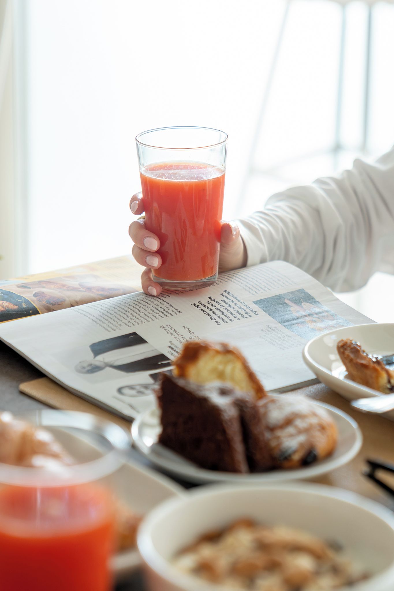 A person holding a glass of red juice, a newspaper, and a plate with a slice of cake.
