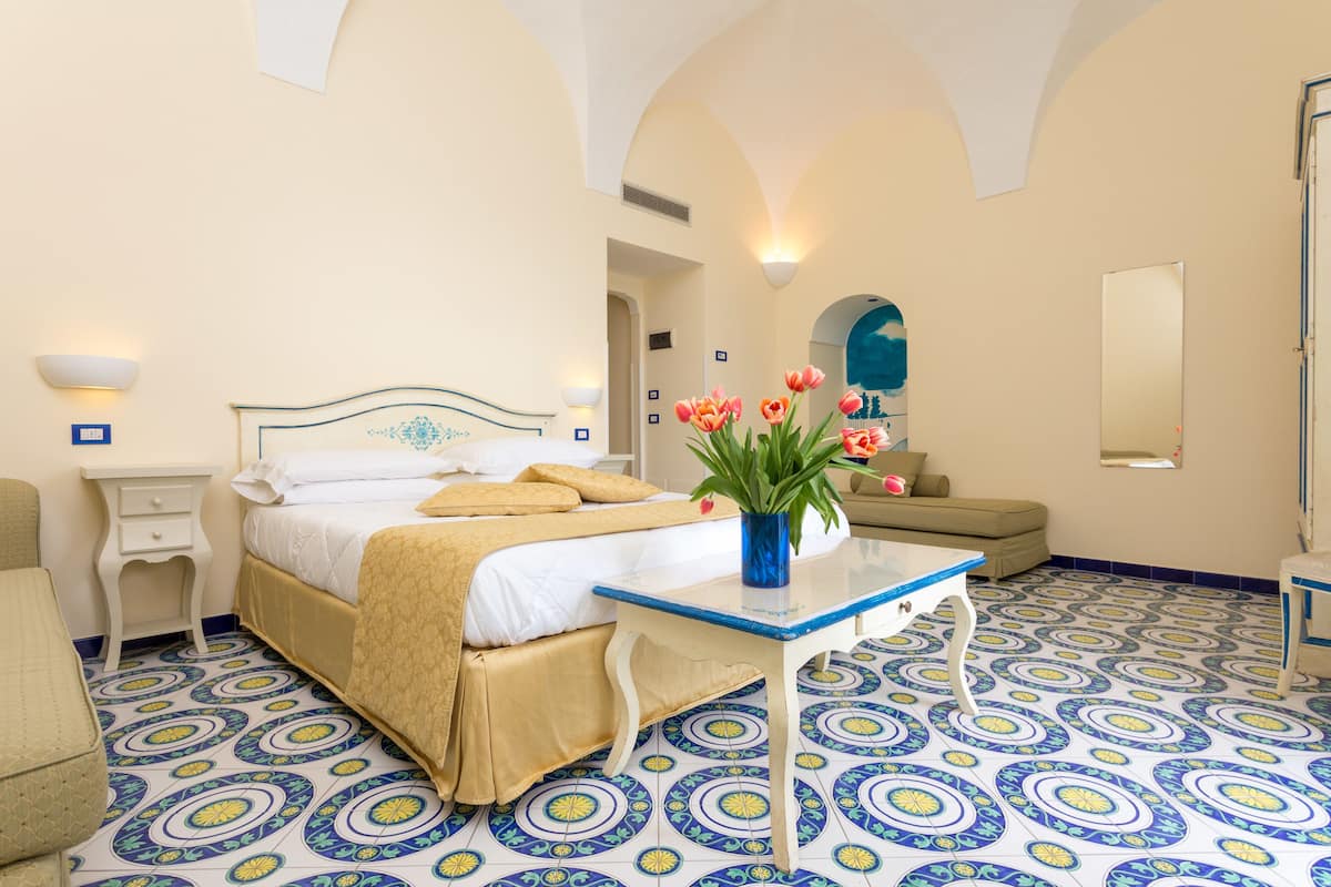 A hotel room with a bed, a bedside table with a vase of flowers, a chair, a sofa, and a mirror.
