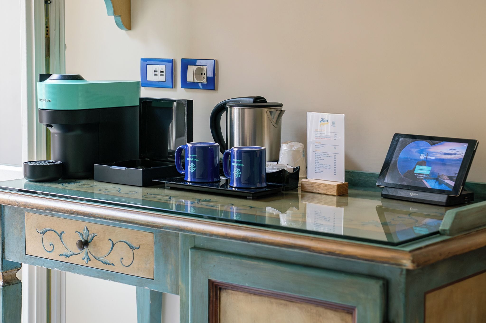 A hotel reception desk with a coffee machine, two blue mugs, a kettle, a menu, and a tablet displaying a scenic view.