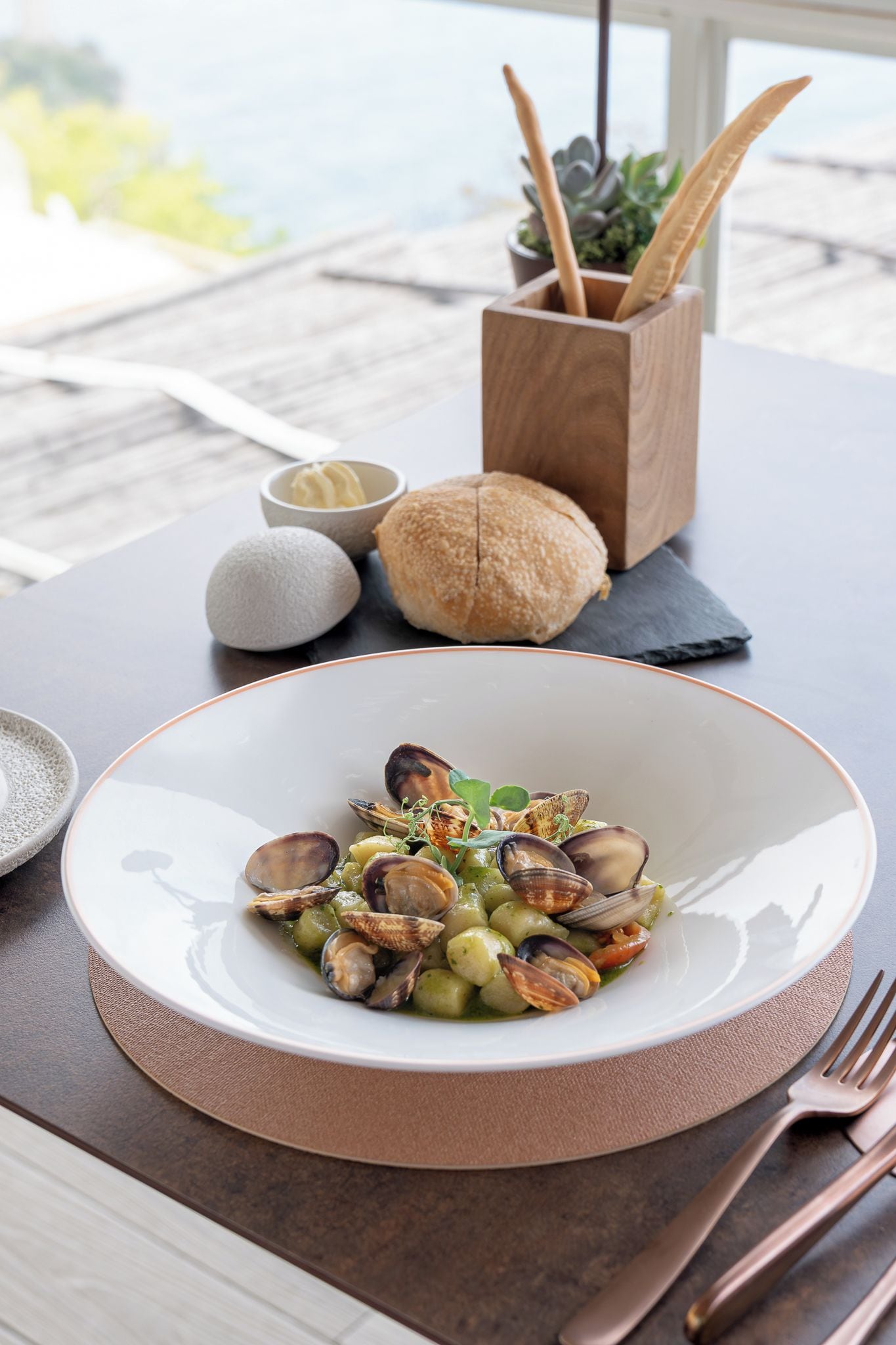 A table with a white plate of mussels, a bowl of butter, a loaf of bread, and a small potted plant.