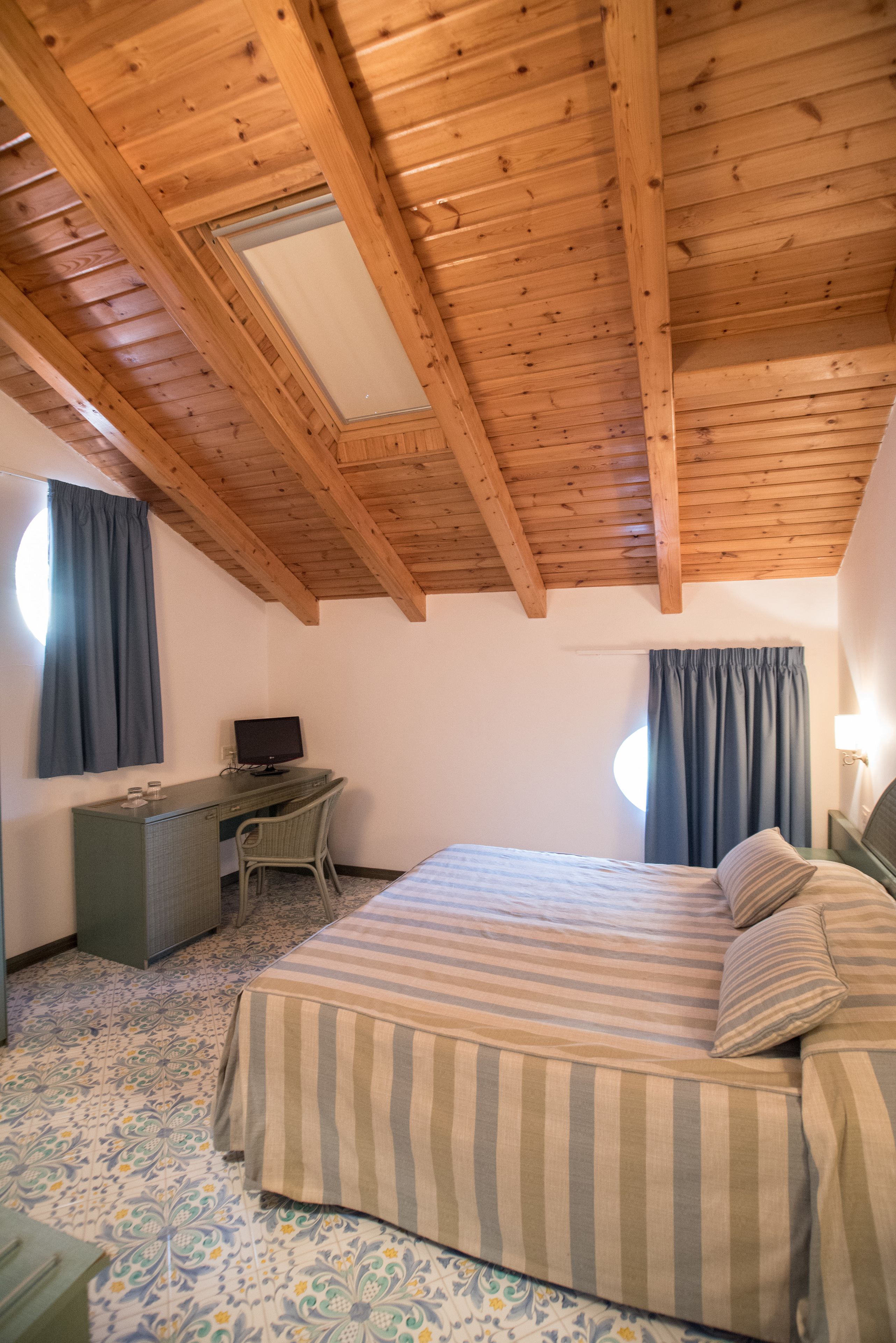 Un dormitorio con techo de madera, una cama con ropa de cama a rayas, un escritorio con una computadora y dos ventanas con cortinas.