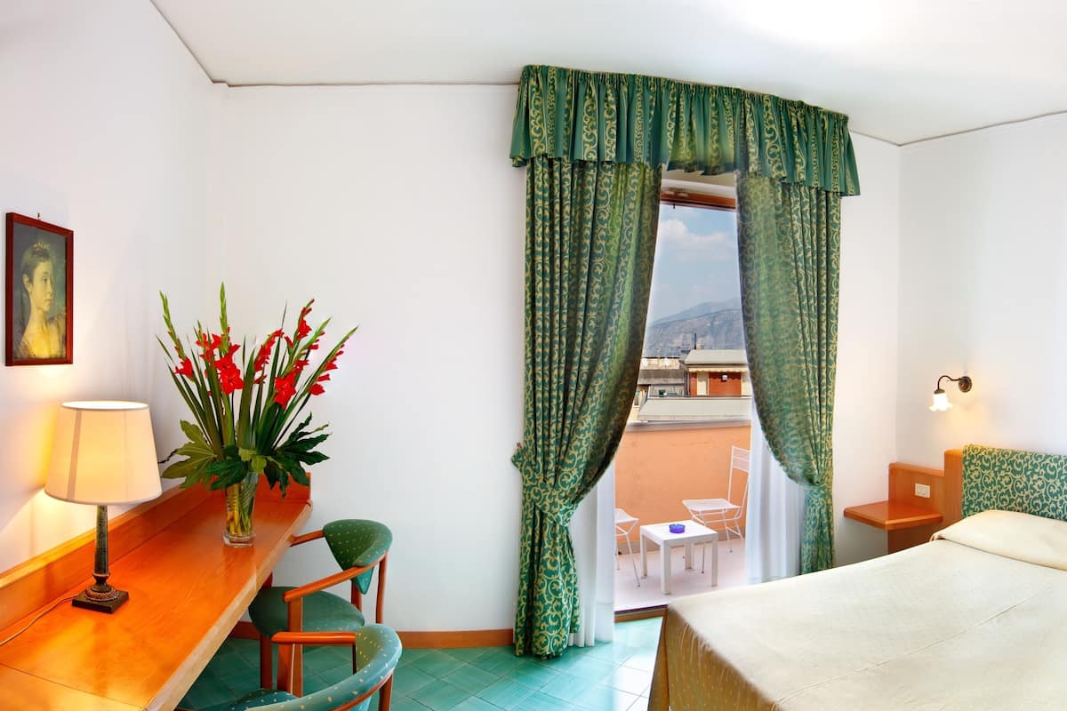 A hotel room with a bed, a desk with a lamp, a chair, a small table, and a view of the city through a window with green curtains.