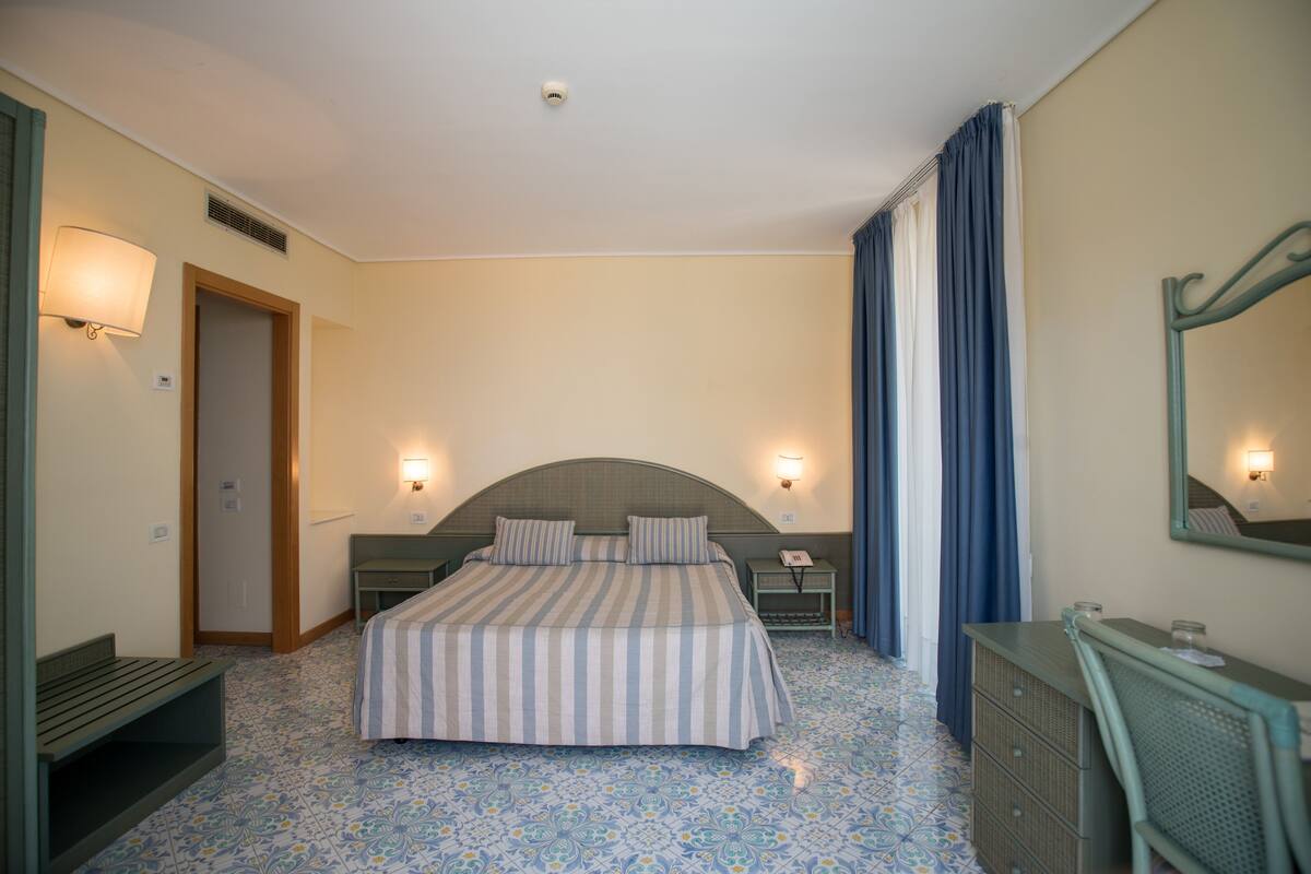 A hotel room with a bed, bedside tables, a desk, a chair, a bench, a mirror, and a telephone.
