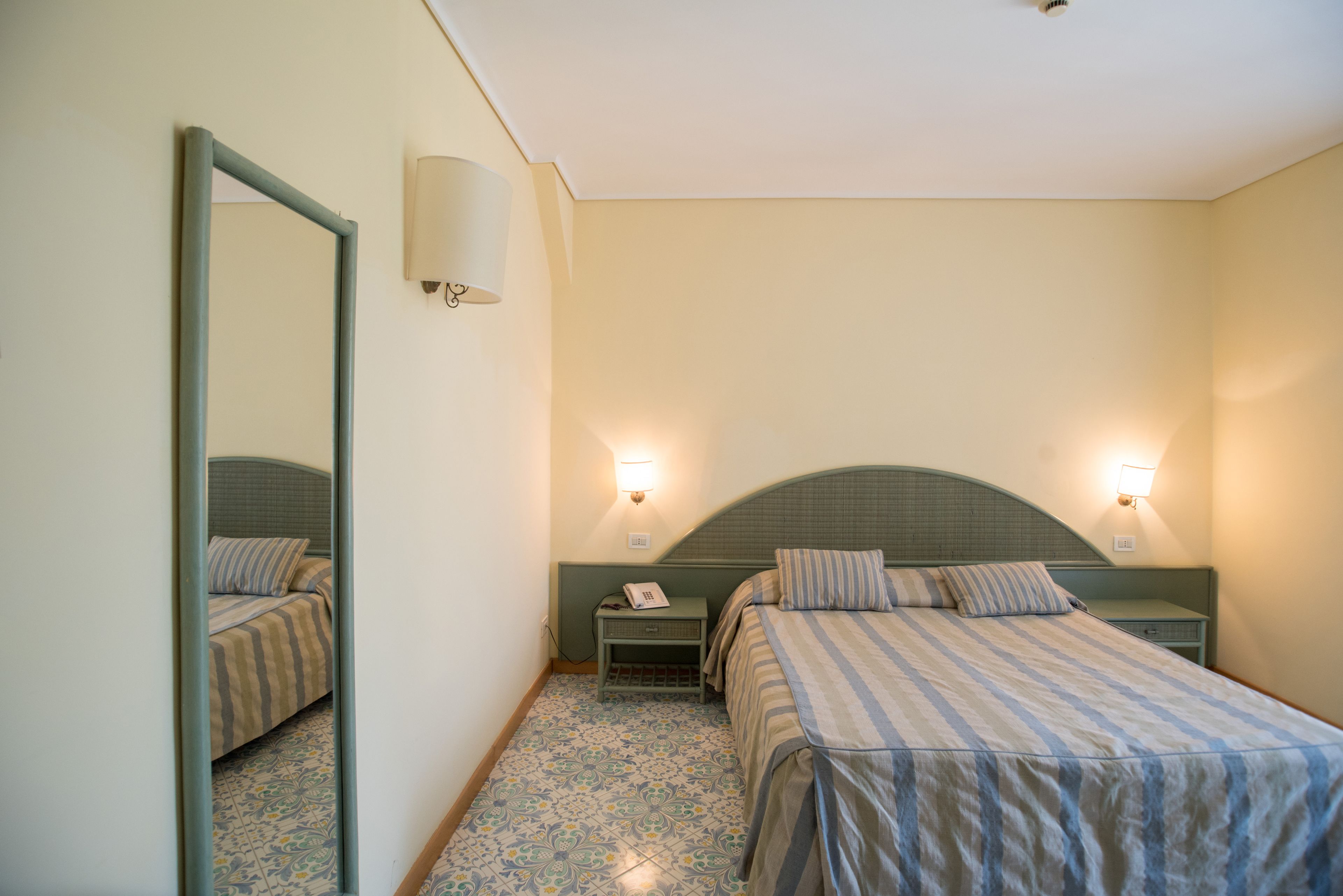 A hotel room with a bed, two bedside tables, a mirror, and a wall-mounted lamp.
