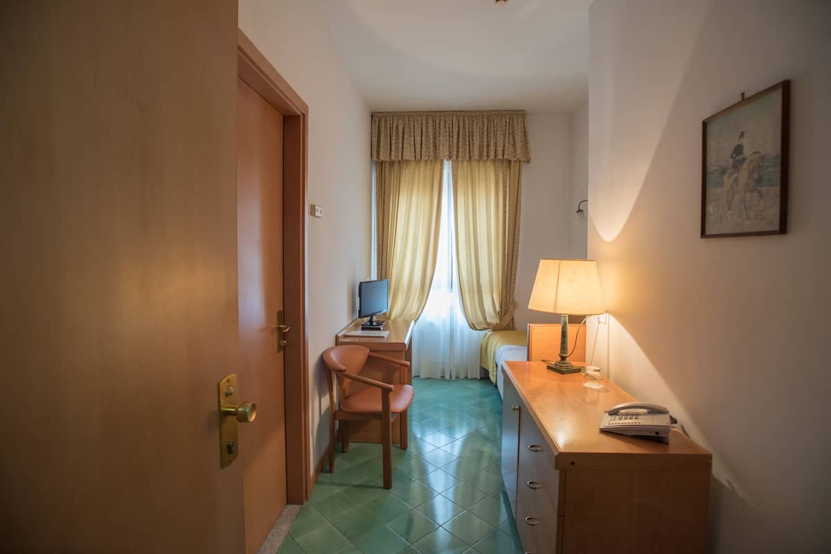 A hotel room with a wooden desk, a chair, a bed with a window, and a lamp.