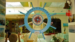 A room with a large ship's wheel as a centerpiece, decorated with various maritime-themed items. - Marconi (Cattolica)