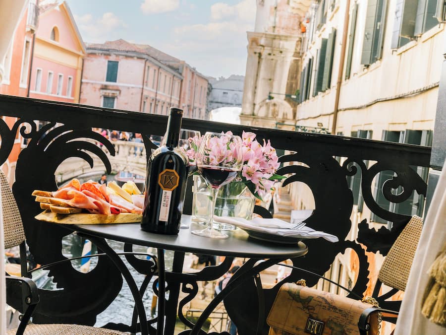 Un balcón con una mesa dispuesta para una comida, que incluye una botella de vino, una copa de vino y un plato de comida.