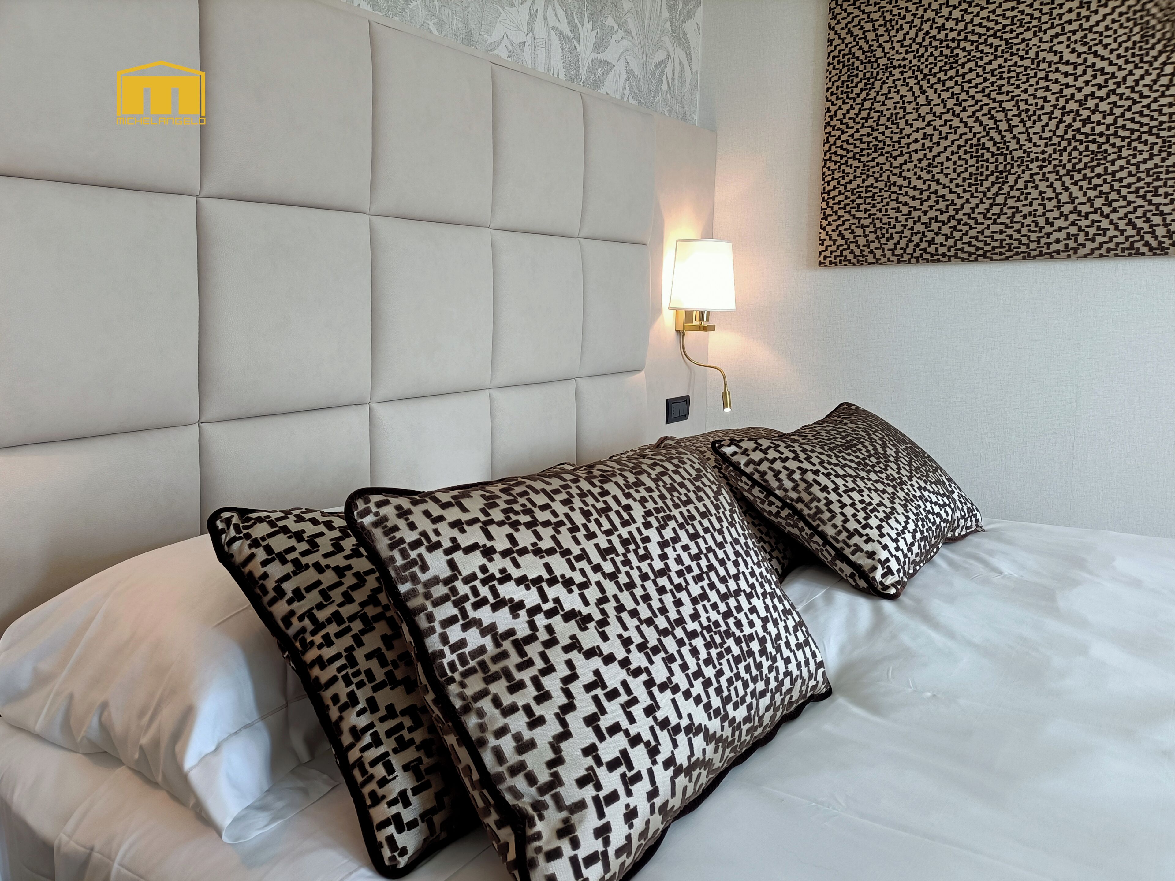 A hotel room with a tufted headboard, patterned pillows, a wall-mounted lamp, and a decorative wall piece.