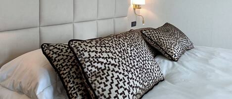 A hotel room with a tufted headboard, patterned pillows, a wall-mounted lamp, and a decorative wall piece.