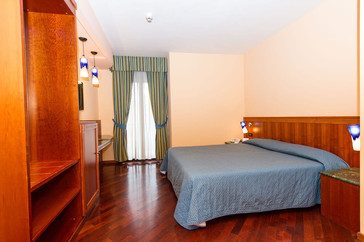 A hotel room with a wooden wardrobe, a bed with a blue bedspread, a wooden headboard, and two wall-mounted lamps.