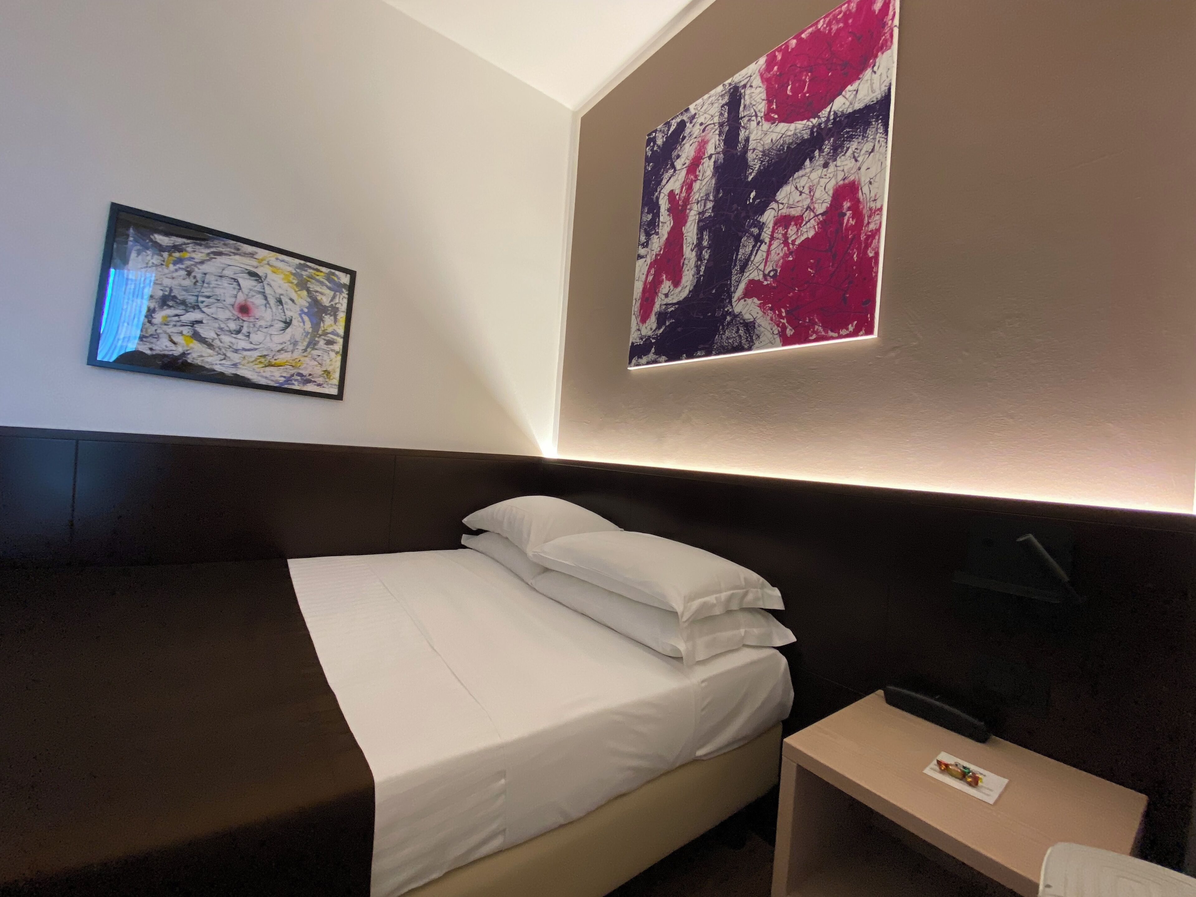 A hotel room with a bed, a bedside table, and two abstract paintings on the walls.