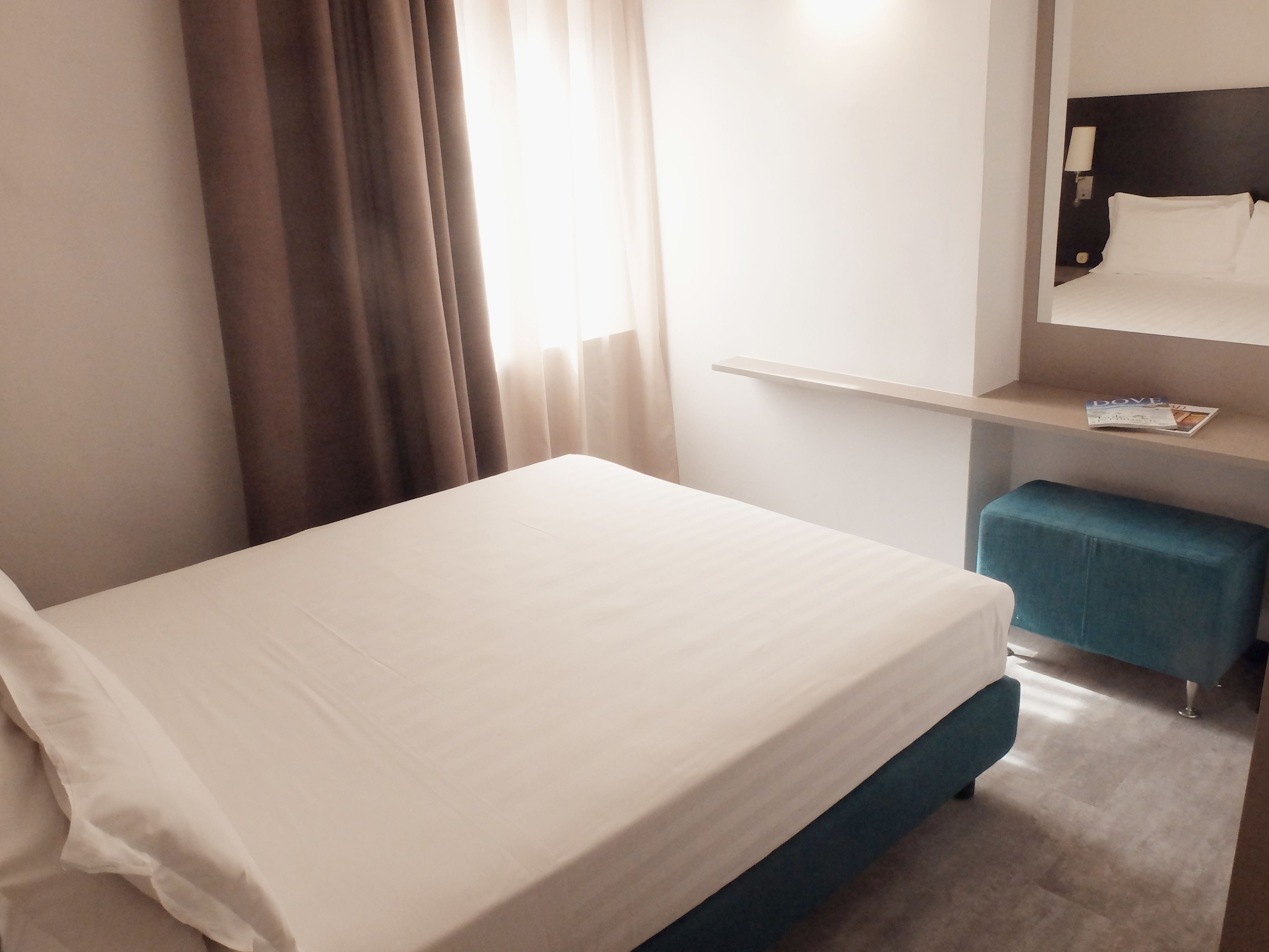 A hotel room with a bed, a nightstand, a mirror, and a shelf with a magazine.
