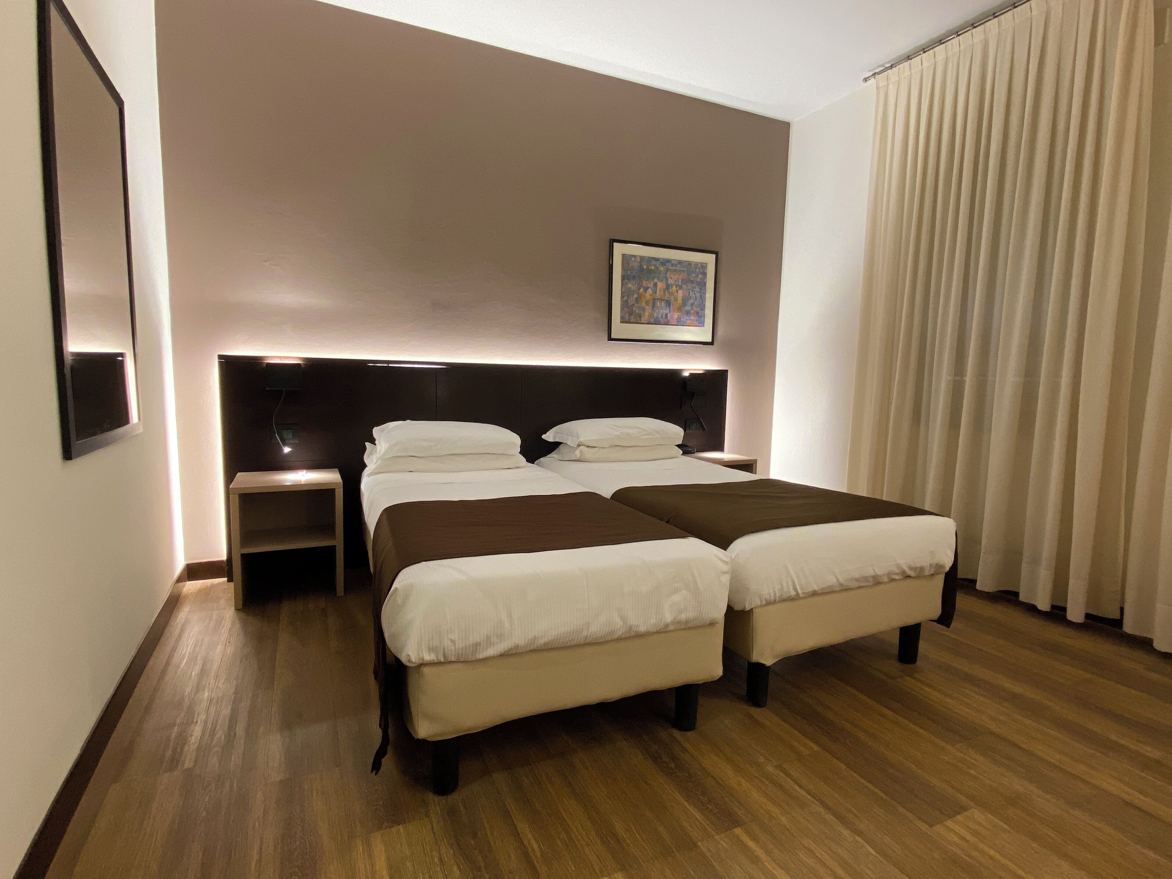 Una habitación de hotel con dos camas, una mesita de noche, un cuadro en la pared y una ventana con cortinas.