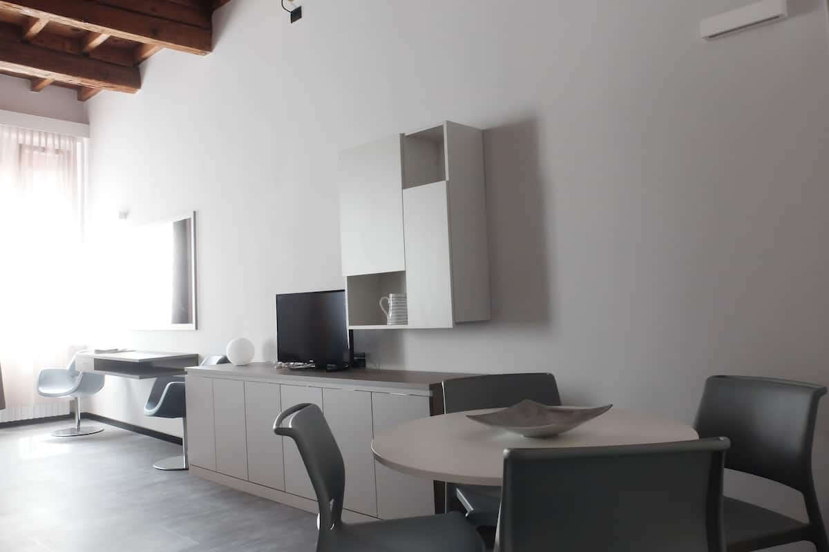 Un comedor moderno con una mesa redonda y cuatro sillas, un televisor de pantalla plana en un mueble bajo, y un amplio ventanal que proporciona luz natural.